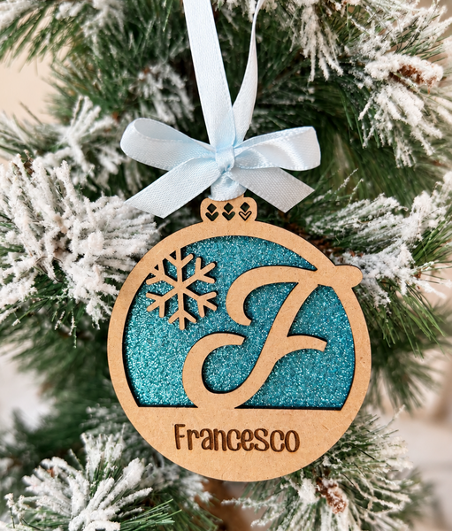 Bola de Navidad de madera con inicial F y nombre Francesco, fondo de purpurina azul, copo de nieve y lazo azul claro, colgada en un árbol nevado.