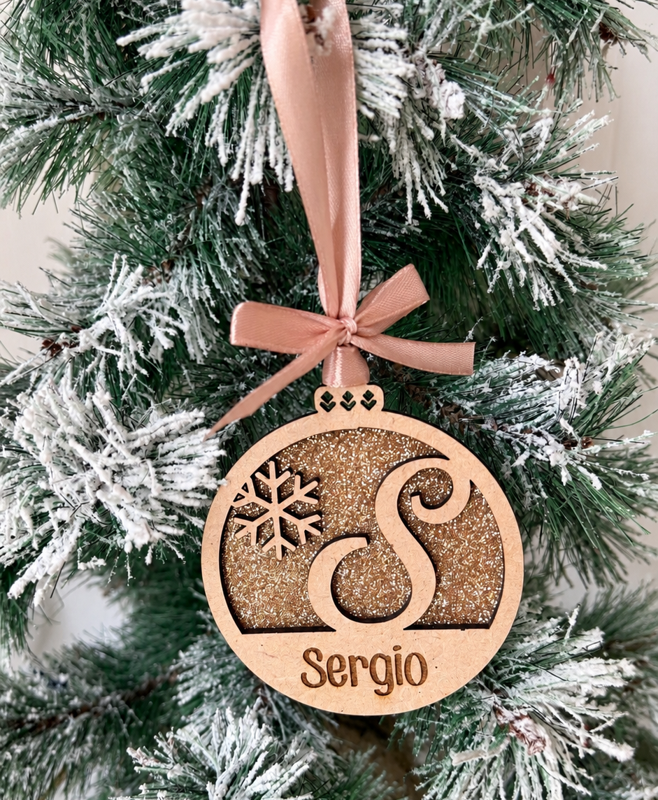 Bola de Navidad de madera con inicial S y nombre Sergio, fondo de purpurina dorada y lazo rosa, colgada en un árbol nevado.