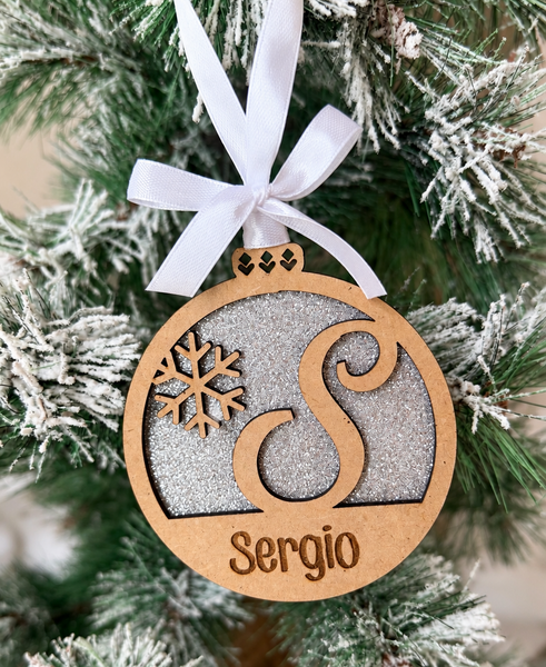Bola de Navidad de madera con inicial S y nombre Sergio, fondo de purpurina plateada, copo de nieve y lazo blanco, colgada en un árbol nevado.