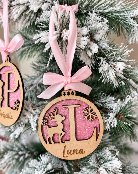 Bola de Navidad de madera con inicial L y nombre Luna, fondo de purpurina rosa, reno, copos de nieve y lazo rosa, colgada en un árbol nevado.