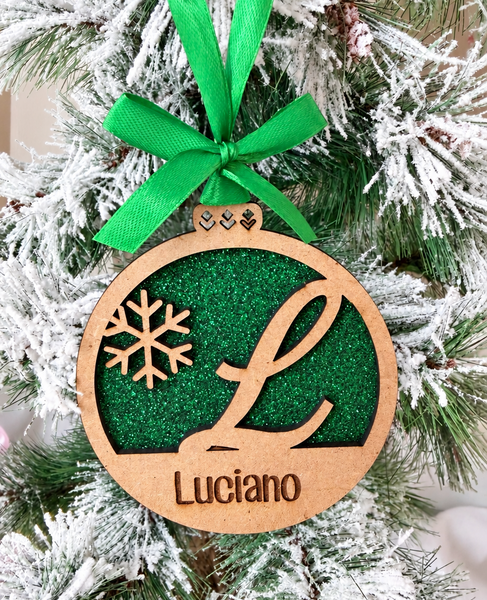 Bola de Navidad de madera personalizada con inicial L y nombre Luciano, fondo de purpurina verde y lazo verde, colgada en un árbol nevado.
